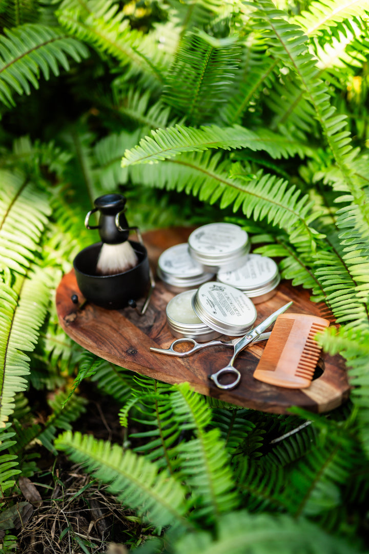 A grooming set arranged on a wooden tray surrounded by lush green fern leaves.