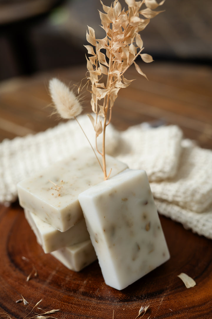 Square soaps stacked on a wooden surface with dried herbs and a white cloth.