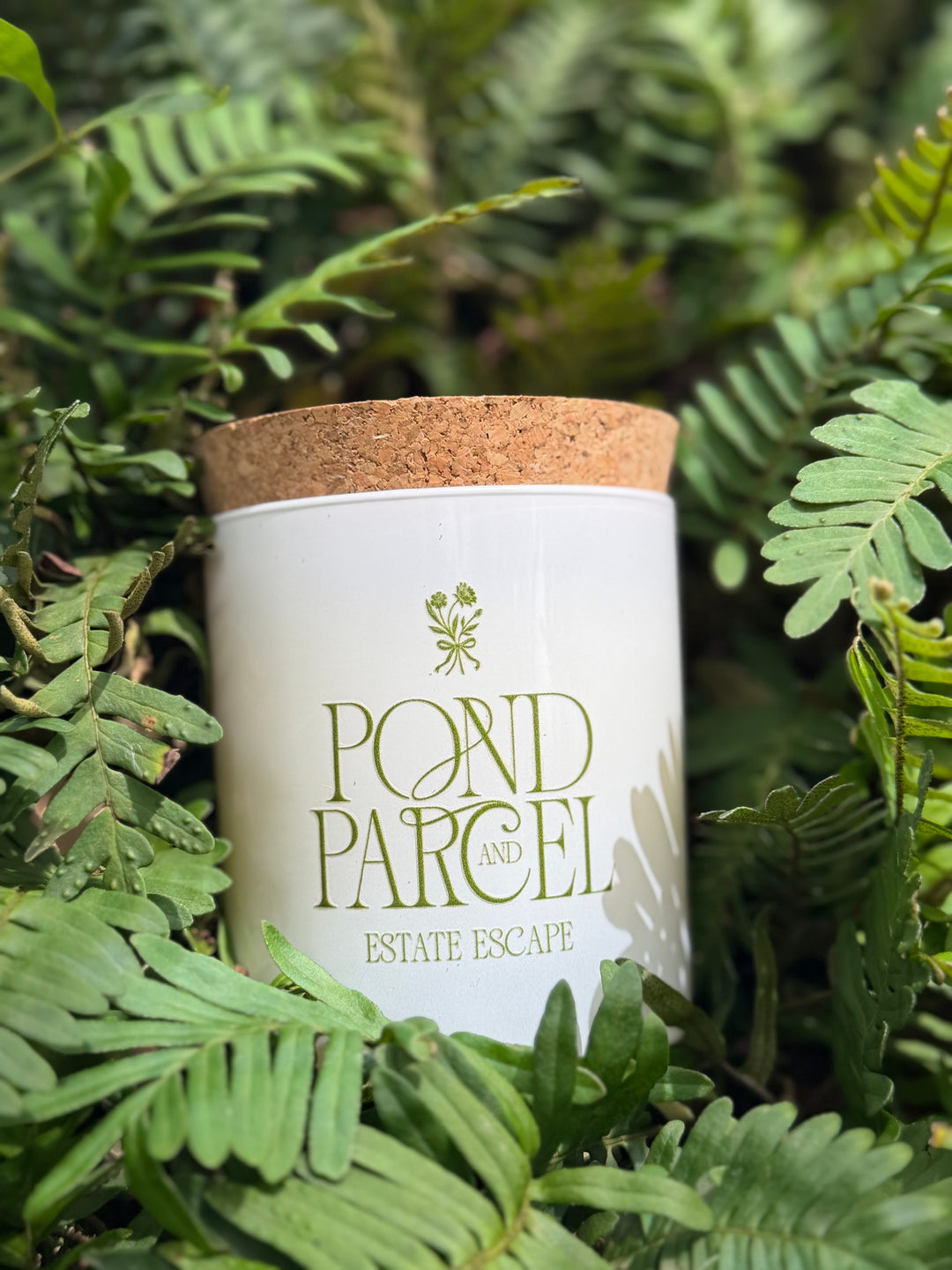 A white candle jar with a cork lid labeled 'POND PARCEL AND ESTATE ESCAPE' in green lettering, nestled among green fern leaves.