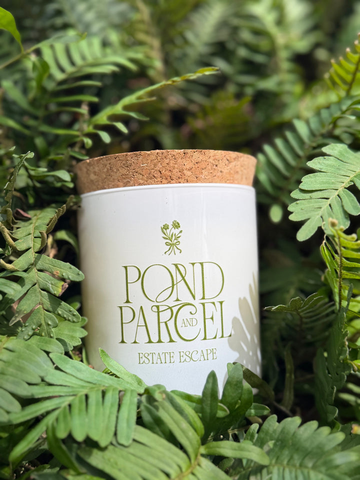 A white candle jar with a cork lid labeled 'POND PARCEL AND ESTATE ESCAPE' in green lettering, nestled among green fern leaves.