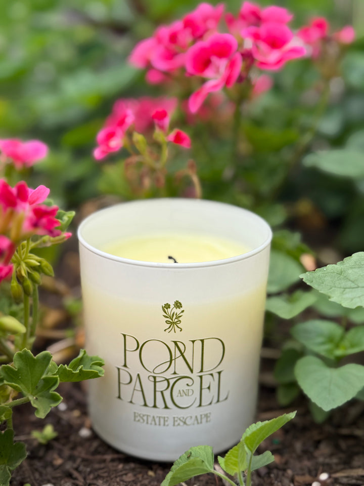 A white glass jar candle labeled 'POND PARCEL AND ESTATE ESCAPE' sits outdoors among green leaves and pink flowers. 