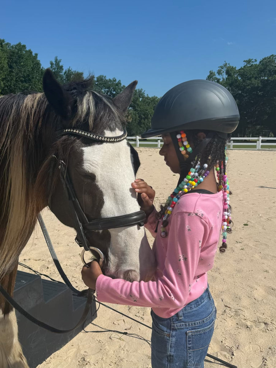 A young girl in equestrian attire, wearing a dark gray riding helmet and a pink long-sleeve shirt, stands in a sandy riding arena next to a brown and white horse, holding its reins. 