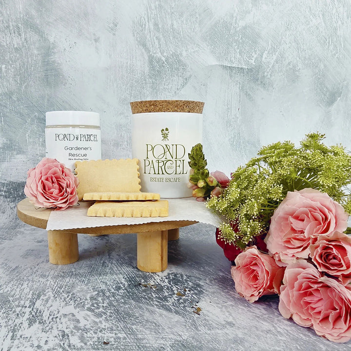 Pond Parcel candles and soap on a wooden stand with pink roses and greenery on a textured surface.