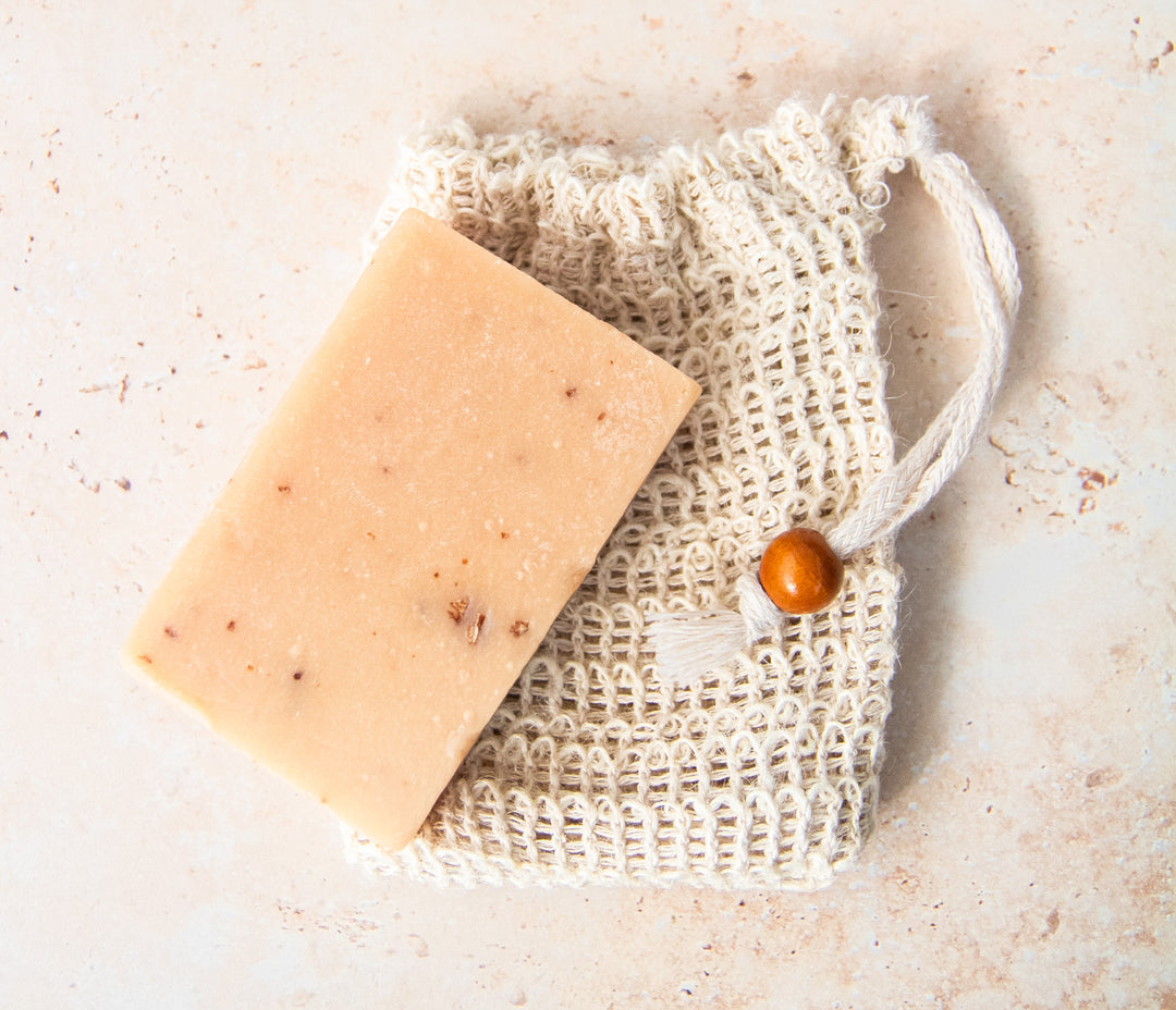 Bar of soap on a natural fiber soap bag with a wooden bead on a light stone background.